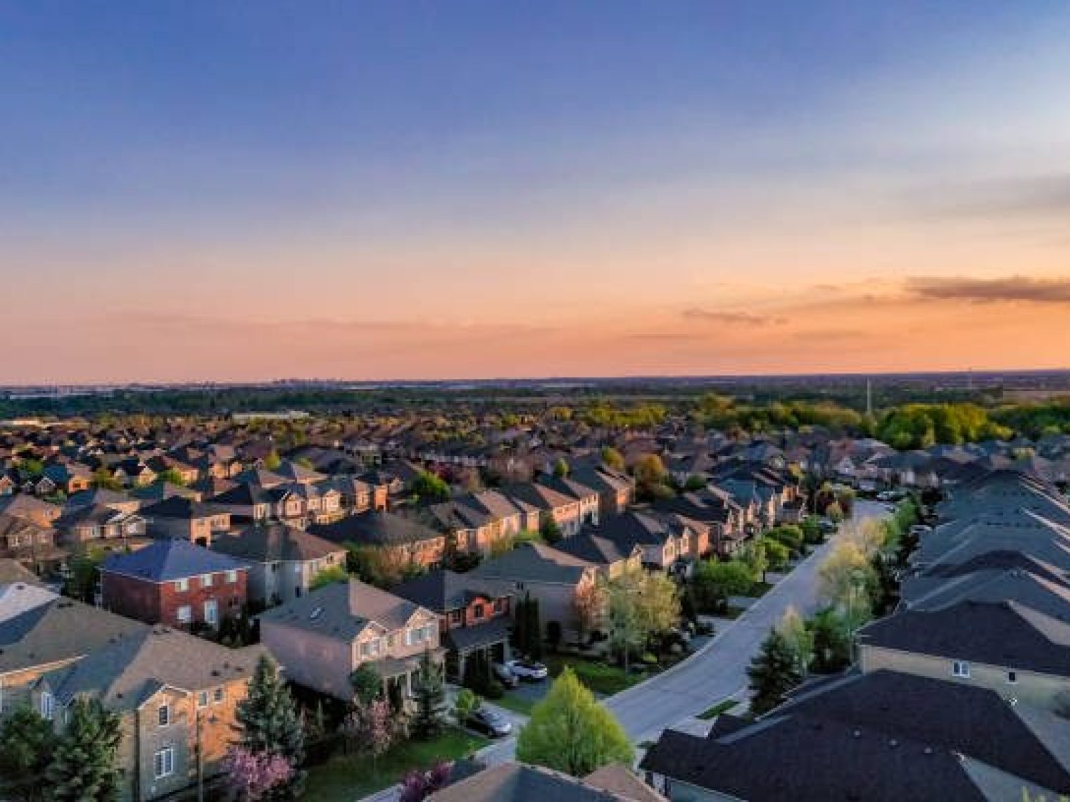 Aerial view of a suburban neighborhood at sunset, featuring rows of houses with rooftops, tree-lined streets, and a colorful sky in the background.