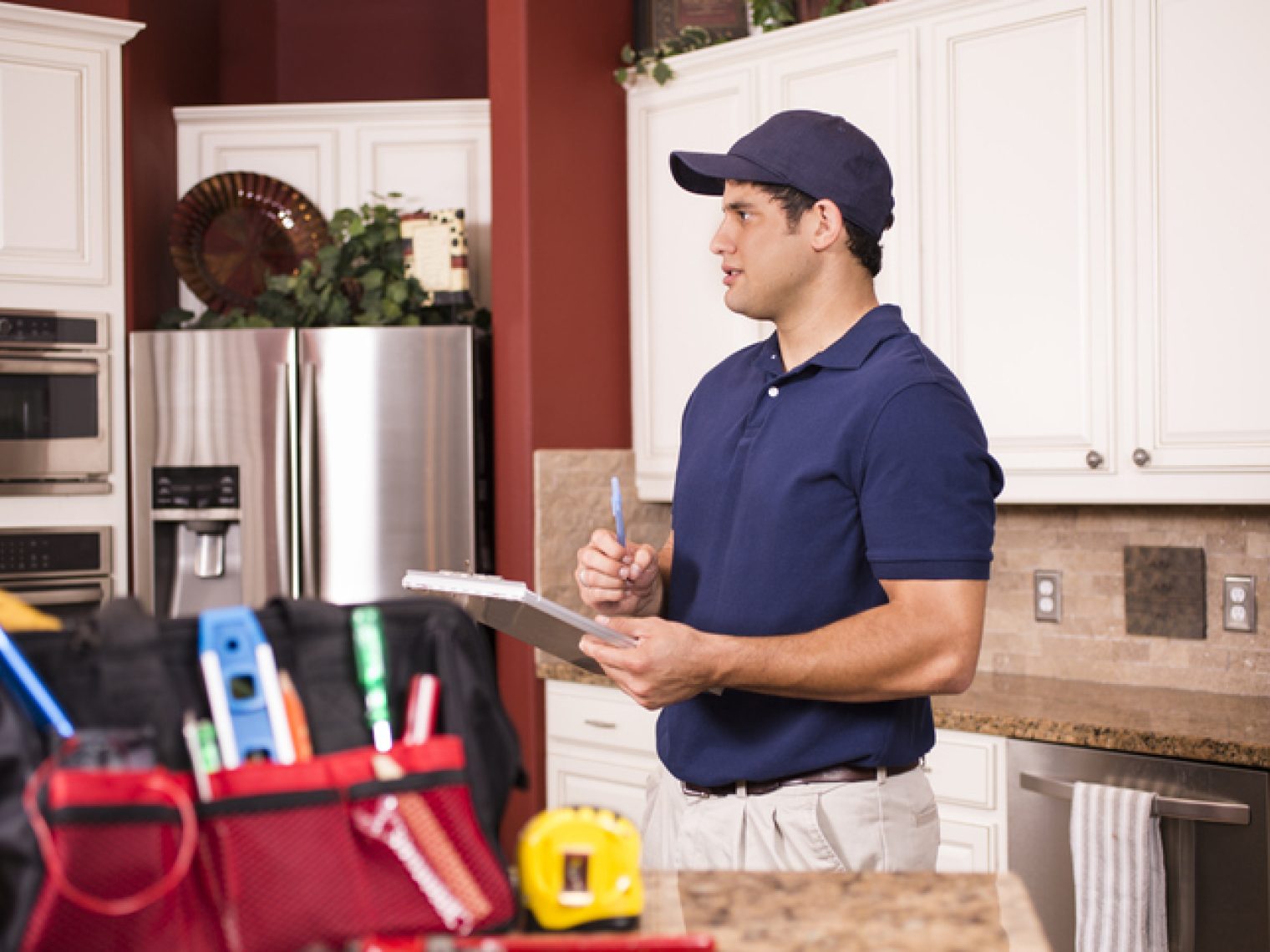 A home appraiser in a blue polo shirt and cap, holding a clipboard, attentively inspects a residential kitchen with a toolkit on the counter.