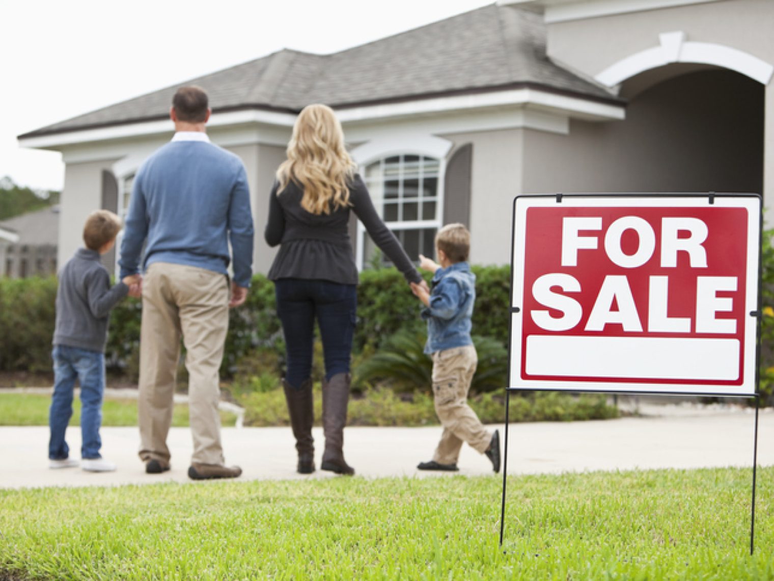 A family of four is seen from behind, walking towards a modern single-story house that is up for sale, indicated by a prominent red "FOR SALE" sign on the well-kept lawn.