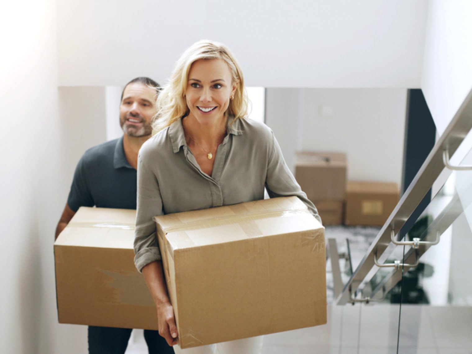 A woman and a man are carrying cardboard boxes up a set of stairs in a bright, modern home, suggesting they are in the process of moving in.