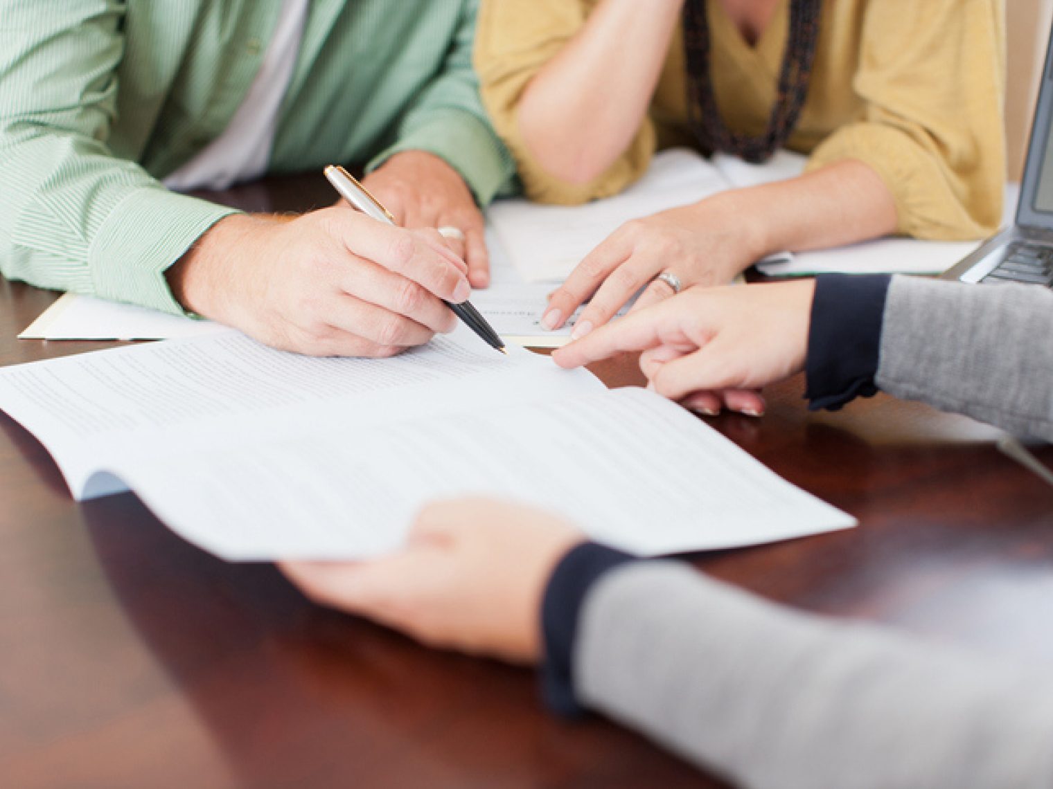 A close-up of three people's hands, with one person pointing at a document while another person writes, with a laptop open beside them on a wooden table.