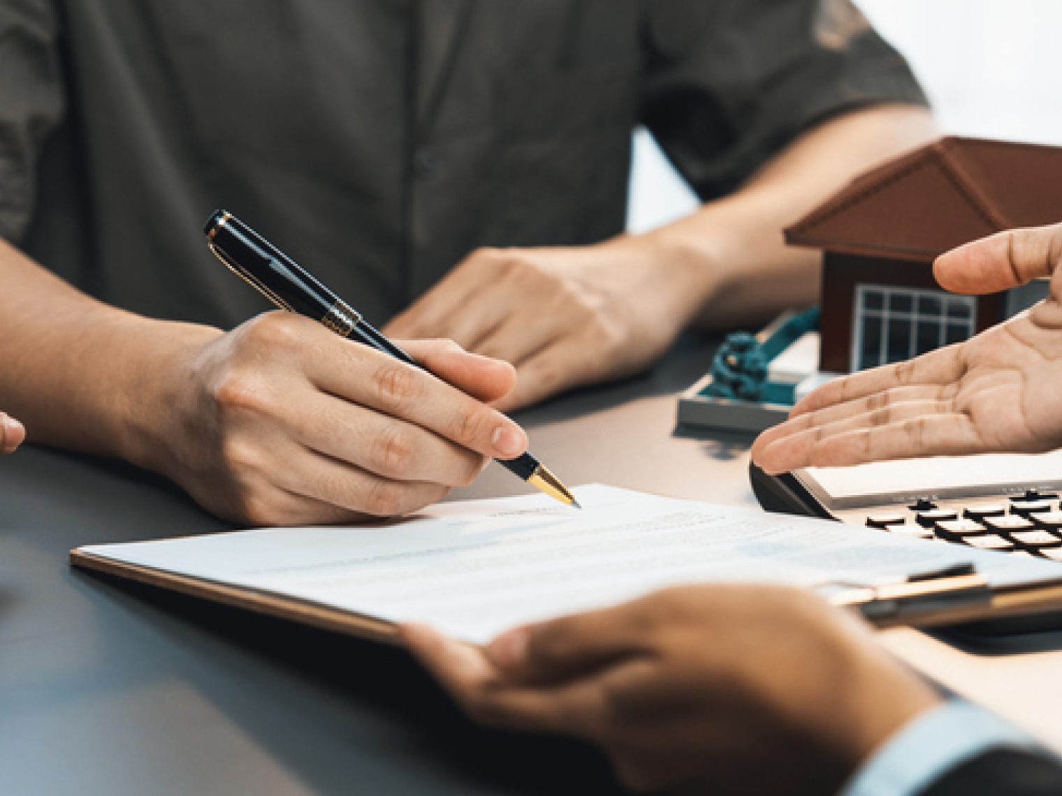 A close-up of a couple's hands on a desk as they are about to sign a document, with a real estate agent gesturing.