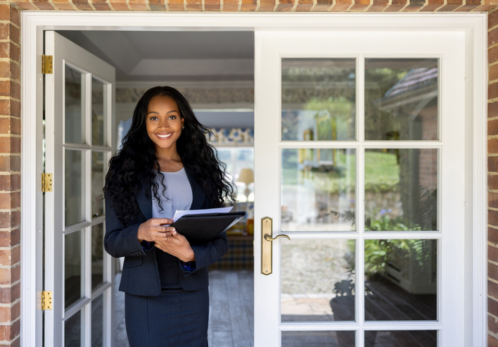 A professional woman with a radiant smile stands at an open door, holding a clipboard, ready to greet or assist, possibly in a real estate context.