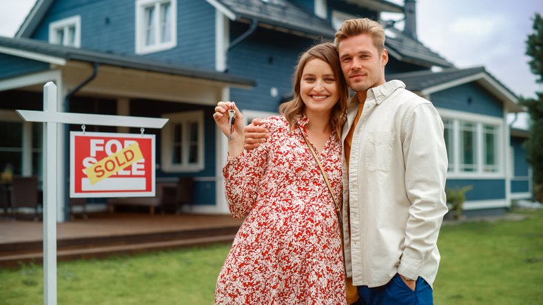 A happy couple embraces and smiles, holding a set of keys in front of a blue house with a "Sold" sign, celebrating their new home purchase.