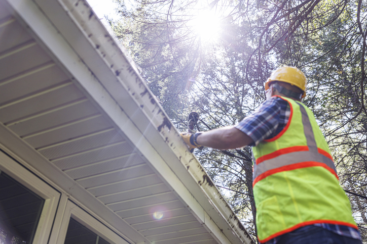 A worker in a high-visibility vest and hard hat repairs or inspects a house's gutter, with sunlight filtering through the trees overhead.