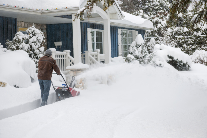 A person in winter clothing uses a snowblower to clear a thick layer of snow in front of a house with snow-covered trees and landscape.