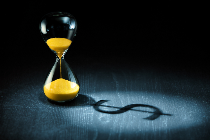 An hourglass with yellow sand on a dark background, casting a long shadow in the shape of a dollar sign.