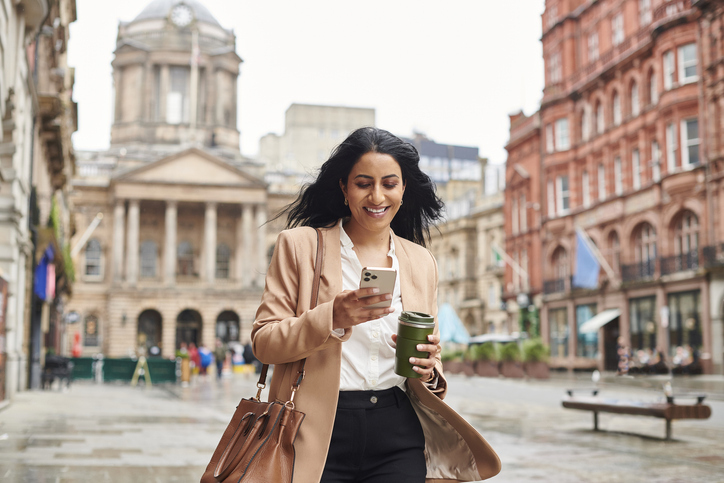 A cheerful woman, walking with a coffee cup and smartphone in hand, looks at her phone screen while walking through an urban square with historic buildings in the background.